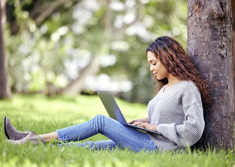 Frau sitzt mit Laptop lehnend an einem Baum im Park